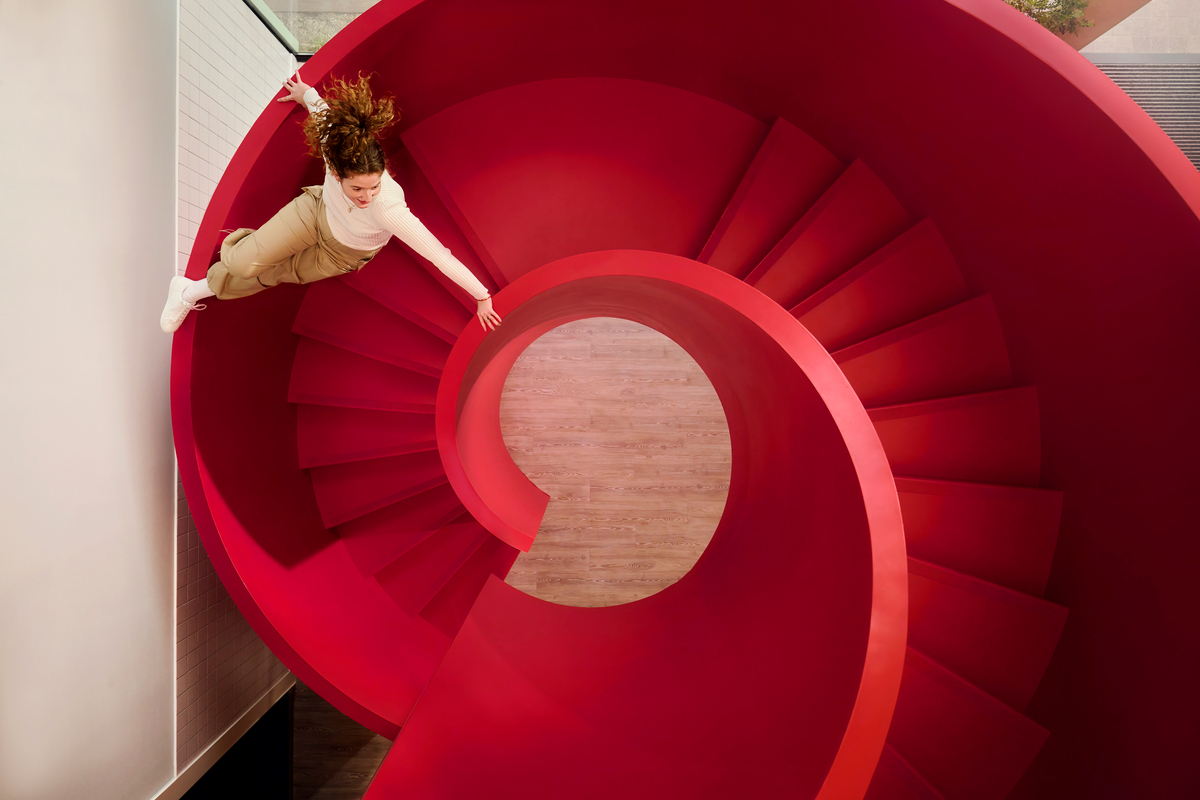 A person in a white top and beige pants playfully leans outward while laying on a vibrant red spiral staircase that twists downward. The background shows a wooden floor.